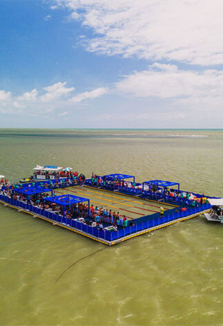 Vista aérea da piscina flutuante do evento AquaRace 2025, instalada sobre uma plataforma modular Pierplas no mar de João Pessoa (PB). A estrutura retangular, cercada por áreas azuis e repleta de espectadores, abriga atletas e equipes durante as provas de natação, com barcos de apoio ao redor.